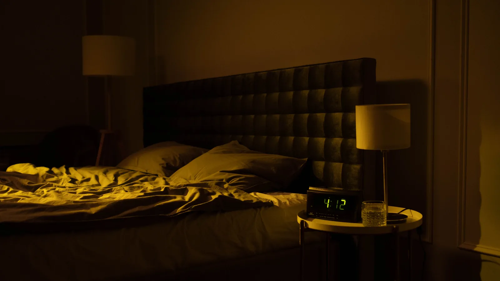 Relaxing dimly lit bedroom with an unmade bed, digital clock displaying 4:12, and warm ambient lighting.