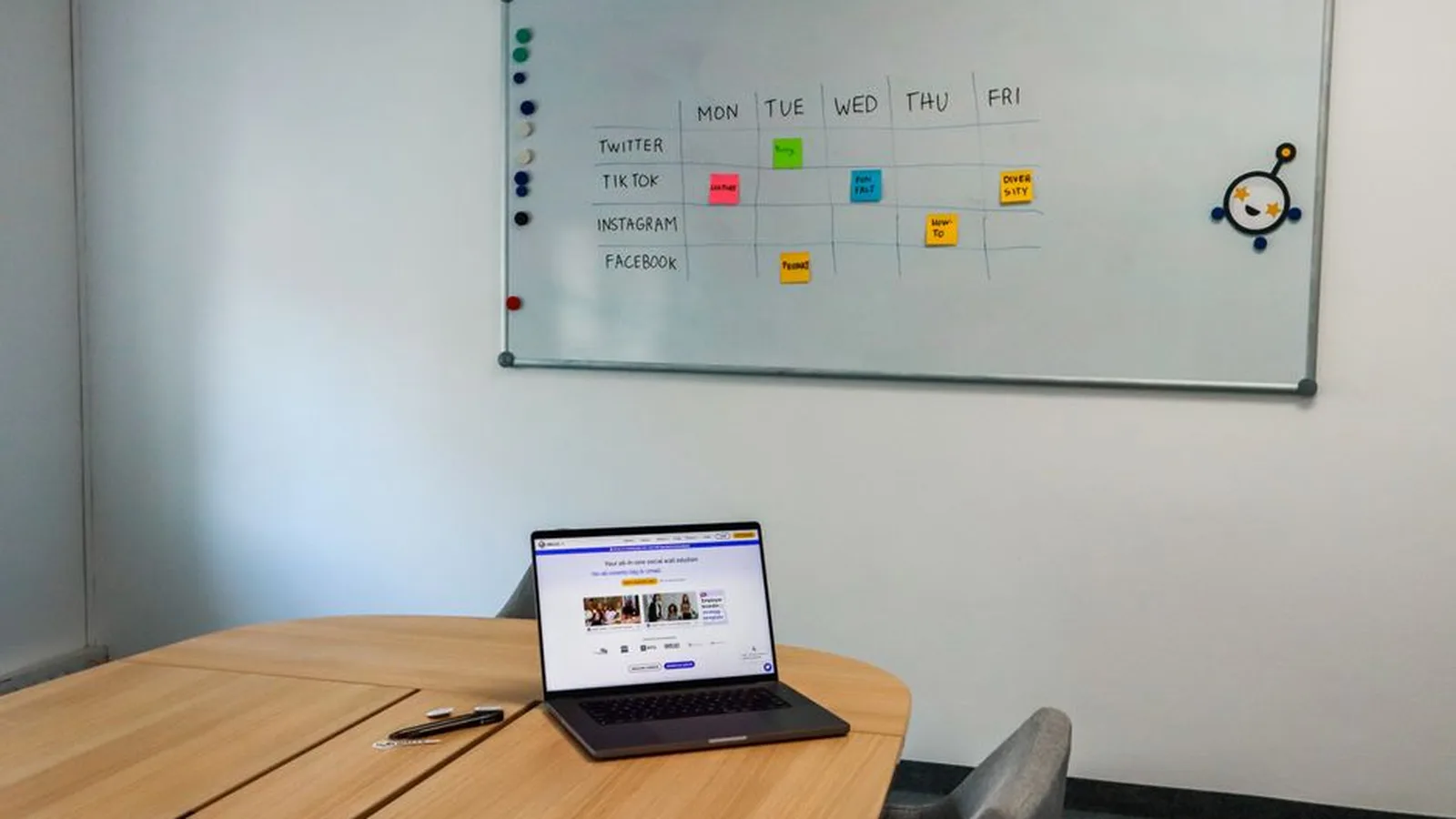 A whiteboard weekly grid listing social apps against Monday through Friday, with a laptop on the table below it.